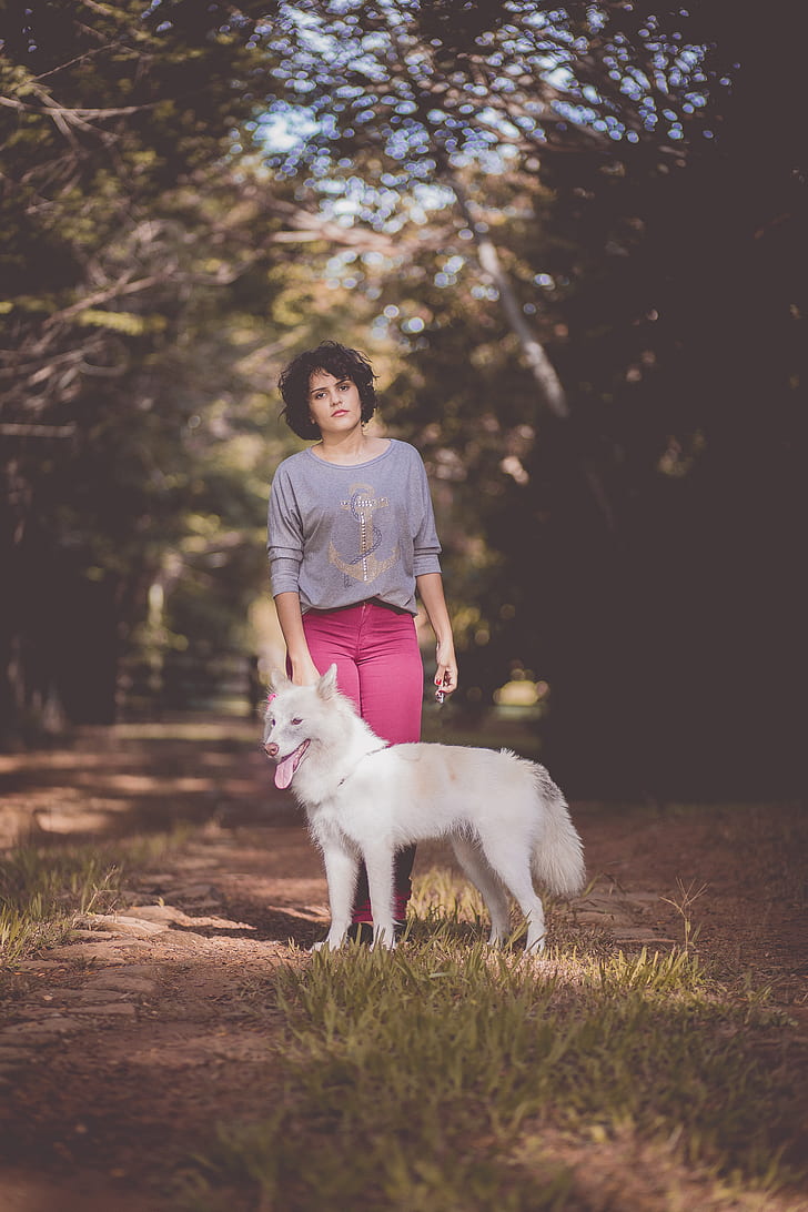 Woman in Purple Long Sleeve Shirt and Pink Jeans Standing Next to White German Spitz With Background of Green Trees during Day