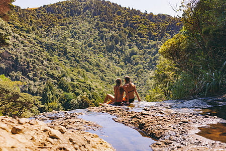 candid photography of two female wearing bikinis sitting on waterfalls cliff in front of green trees
