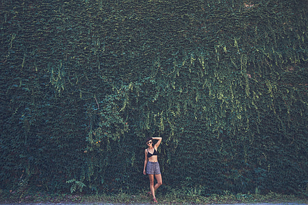photo of woman wearing black bikini top and gray shorts standing near plant wall