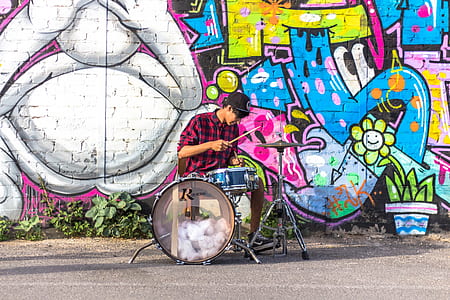 man wearing black and red checkered long-sleeved collared shirt playing drums near graffiti painted wall during daytime