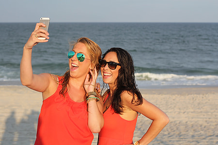 two woman wearing red tank tops taking photos on white sand beach shore