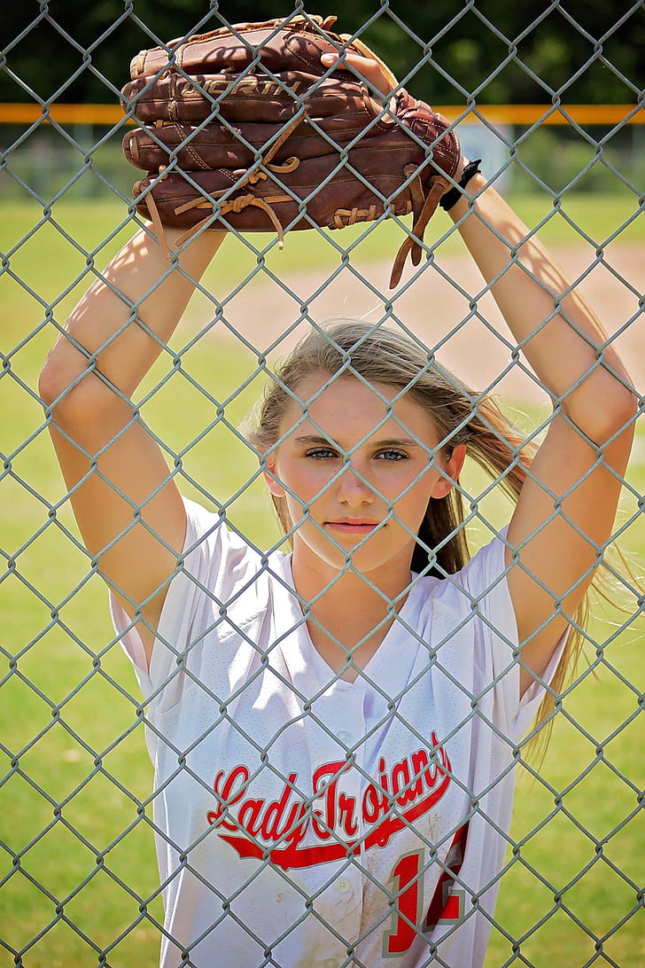 woman wearing white and red Lady Trojant button-up jersey and brown baseball mitt leaning on gray chain link fence