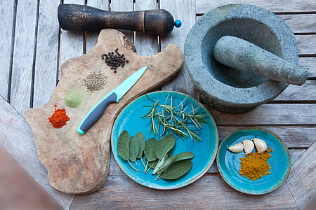 gray mortar and pestle next to blue saucer with garlic and brown powder next to blue plate with green leafs next to brown chopping board with knife and four seasoning powders
