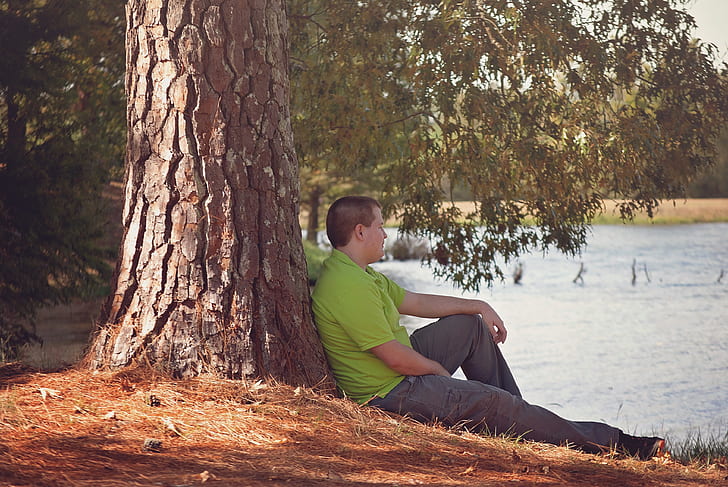 man wearing green shirt sitting lean on tree beside body of water