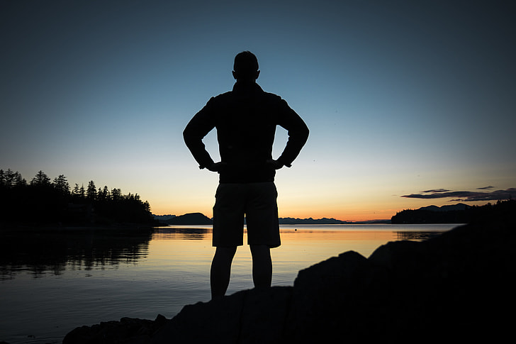 silhouette of man standing on stone near the calm body of water during sunrise
