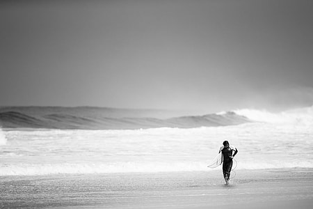 gray scale photography of man holding surfboard on seashore
