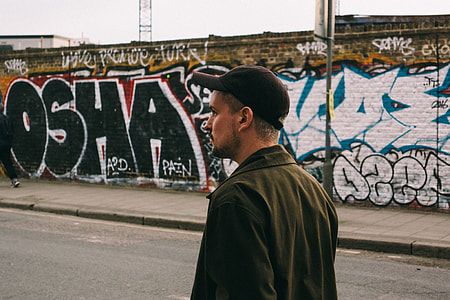 man wearing black jacket and cap walking on street with graffiti wall
