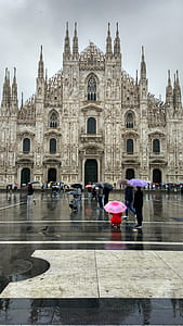 group of people holding assorted-color umbrellas standing near beige concrete cathedral during cloudy skies