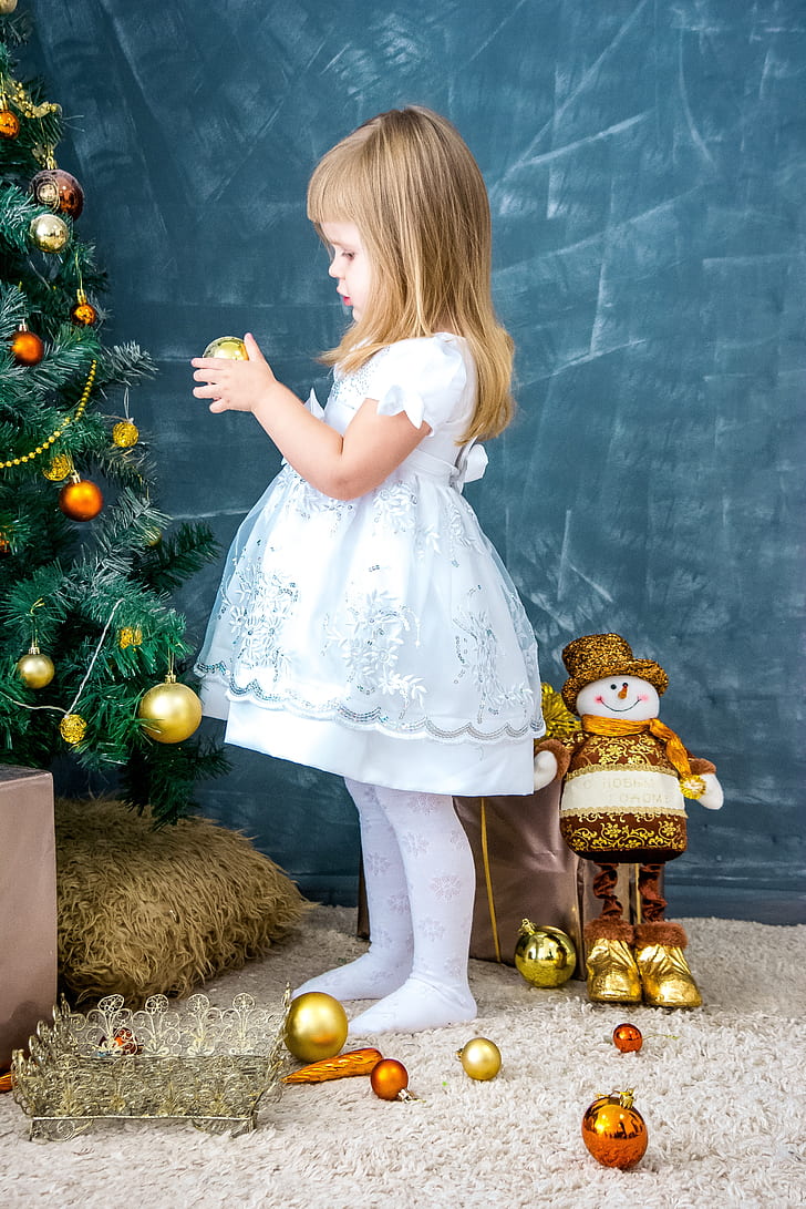 girl in white and light-blue dress holding gold-colored Christmas bauble