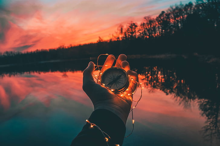 person holding compass and string light near body of water during sunset