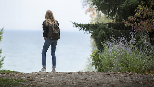 woman in black jacket and blue jeans wearing backpack standing near plants