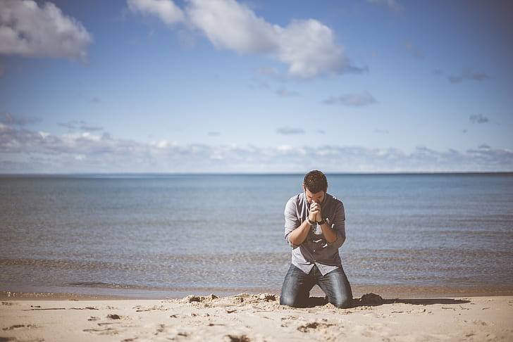 man wearing grey shirt kneeling down on beach shore praying under cloudy sky