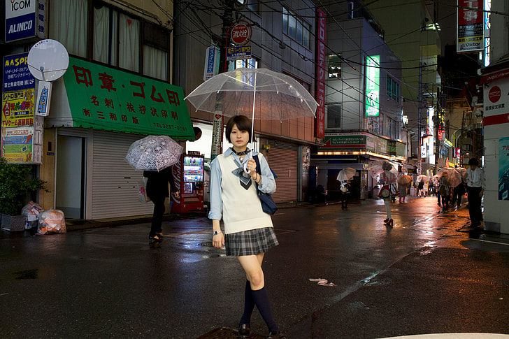 woman holding clear umbrella wearing white and blue collared long-sleeved shirt with black and gray plaid mini skirt standing surrounded by buildings during night
