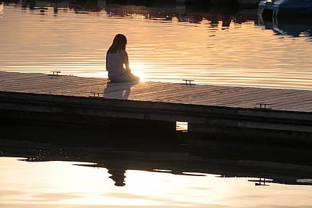 woman in white long-sleeved shirt sits on brown wooden dock