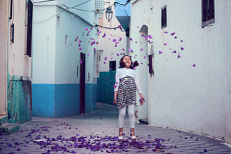 girl in white and black striped long-sleeved shirt and white pants standing in front of white painted wall with purple petals