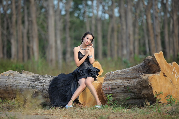 woman wearing black strapless cocktail dress sitting on tree log during daytime in selective-focus photography