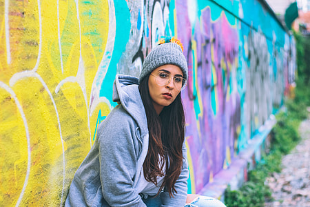 girl in gray jacket and bobble hat sitting on concrete wall with bench and painted graffiti at day time