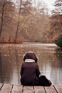 person in black coat sitting on wood above body of water