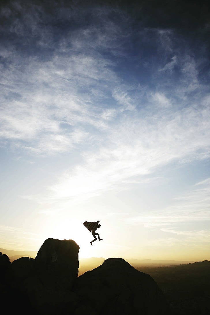 silhouette of person jumping on rock formation under white cloud blue skies during sunrise
