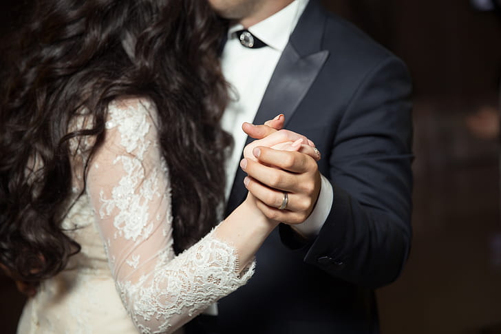 man wearing black peak lapel suit jacket and woman wearing white lace long-sleeved dress holding hands while dancing
