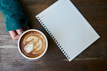person holding white mug with coffee beside notebook