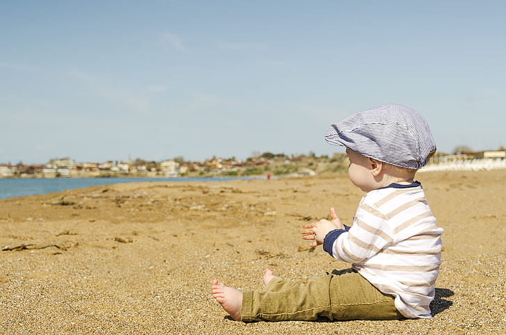 baby in white and brown striped long-sleeved shirt and pants outfit sitting on brown sand at daytime