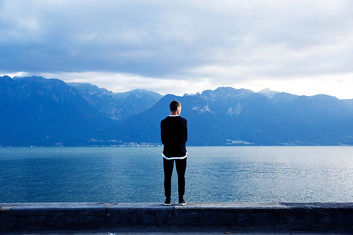 man in black sweater facing the alps across the calm sea during daytime