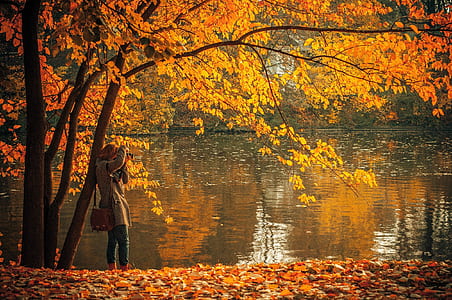 photo of woman in brown suit standing near tree and body of water during daytime