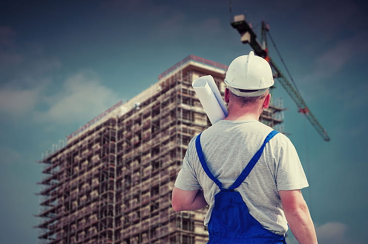 man in white hard hat, gray crew-neck t-shirt and blue jumpsuit holding paper watching building