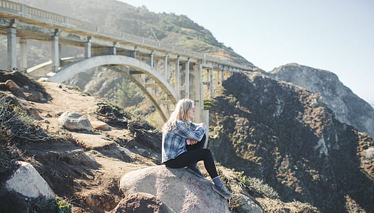 woman in dress shirt sits on grey rock with bridge background
