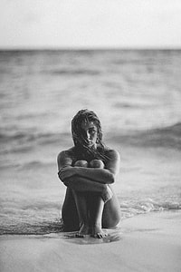 grayscale photo of  female sitting on sand beside seashore
