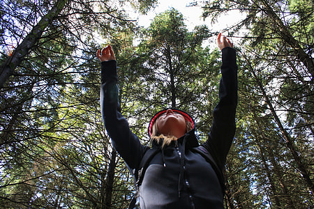 low angle photo of woman wearing black zip-up hooded jacket raising her two hands above with green leaf trees at daytime