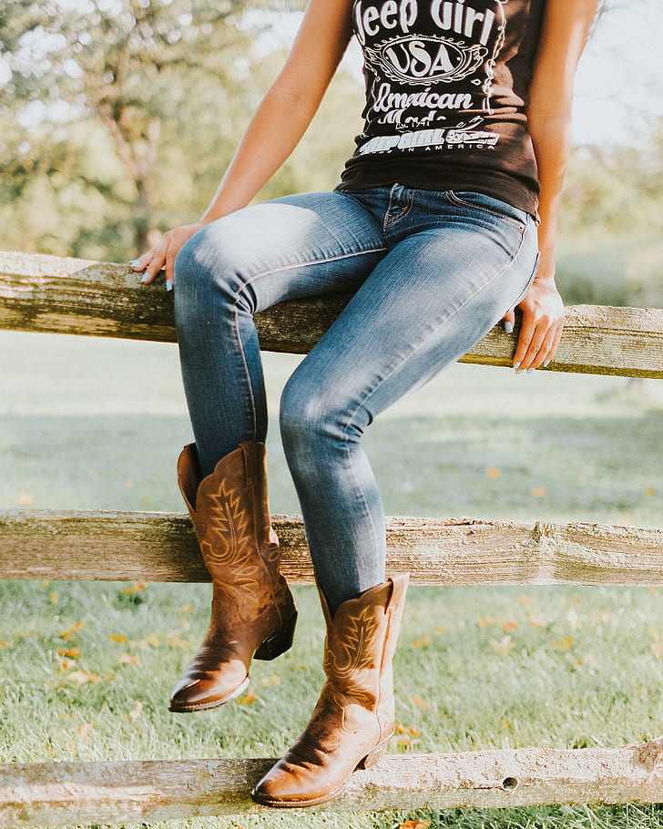woman wearing brown cowboy boots sitting on brown wooden fence