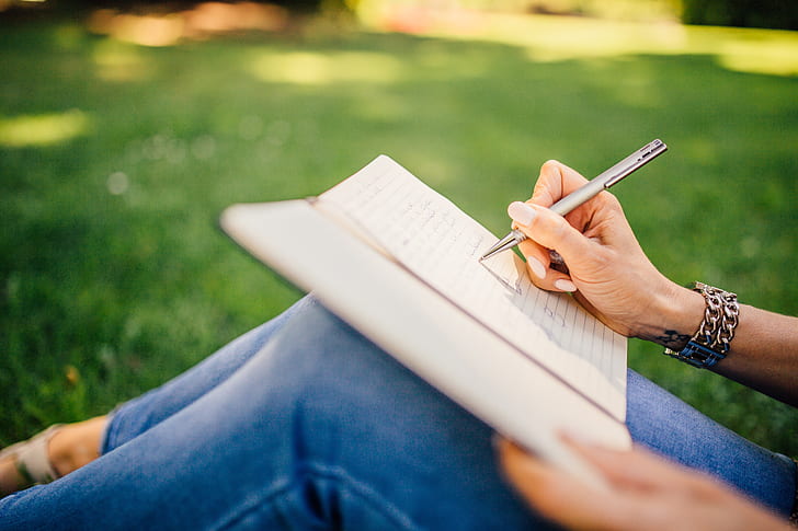 person writing on notepad sitting on green grass in selective focus photography at daytime