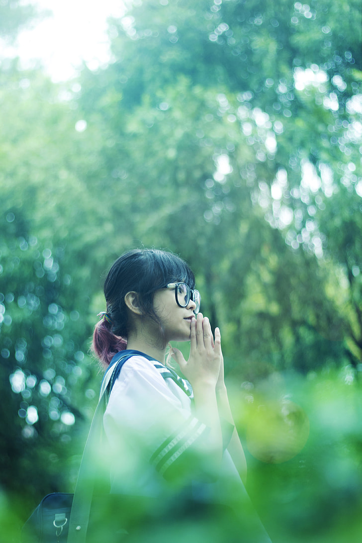 shallow focus photography of black haired girl wearing eyeglasses surrounded by green trees