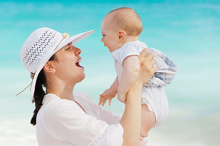 woman in white top and white sunhat carrying baby in white onesie on seashore at daytime