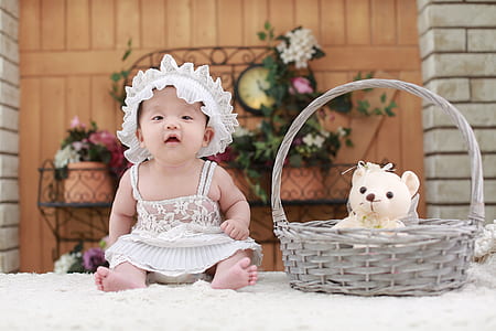 baby wearing white floral dress sitting beside gray basket with white teddy bear inside on fleece mat