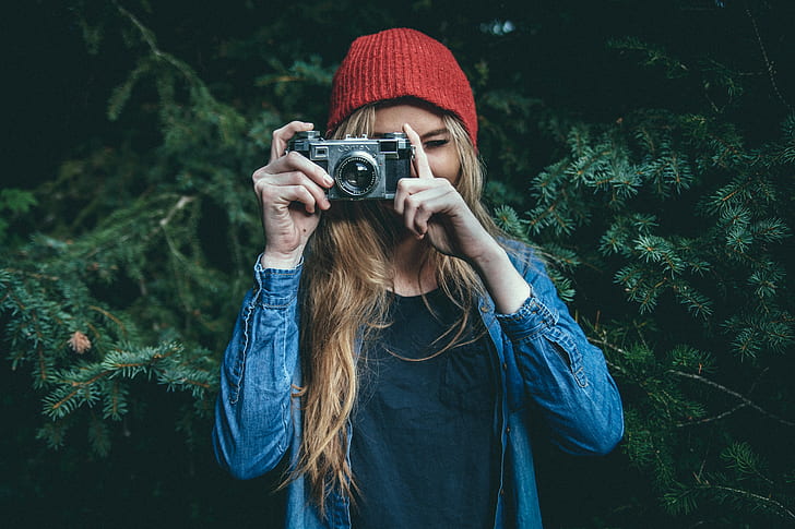 woman wearing blue denim jacket and red beanie taking photo with grey SLR camera near green-leafed tree