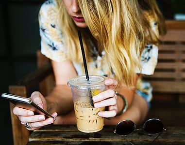 woman wearing white and multicolored floral shirt sitting on brown bench holding disposable cup and using smartphone