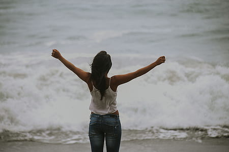 woman wearing white spaghetti strap top and blue bottoms raising hands facing ocean waves