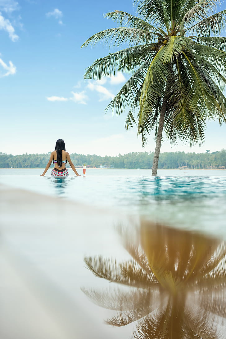 woman in bikini sitting on pool near green coconut tree during daytime