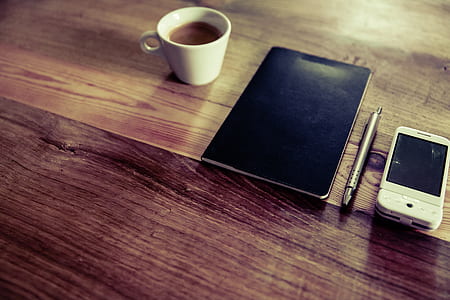 black notebook near click pen, white phone and white ceramic teacup with coffee