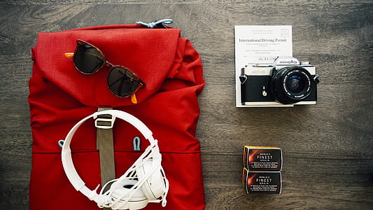 white full size corded headphones, sunglasses on red leather backpack beside gray SLR camera