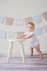 baby wearing white footsie holding into white wooden stool