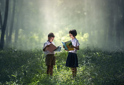 girl and boy holding book while standing in grass field