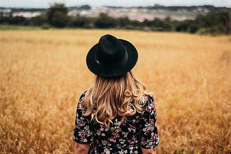woman in black and multicolored floral short-sleeved top and black fedora hat standing on dried grass field