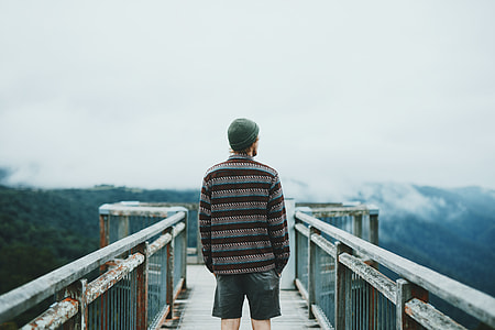 man wearing brown-and-grey striped sweater, blue denim jeans, and green cap standing in the middle of metal bridge overlooking view full of trees under cloudy sky during daytime