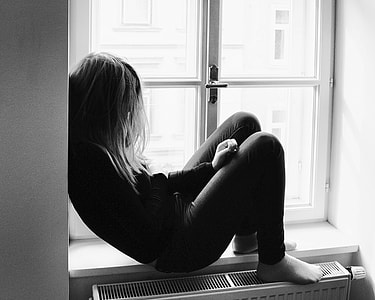 grayscale photo of woman sitting in the window