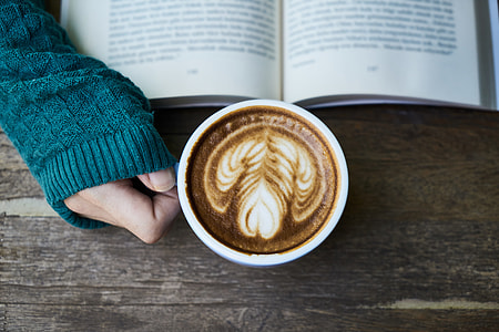 person holding mug of cappuccino next to book