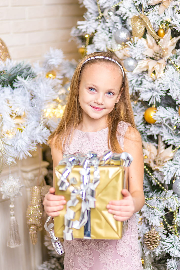 girl in pink lace sleeveless dress holding gold gift box in front of Christmas tree